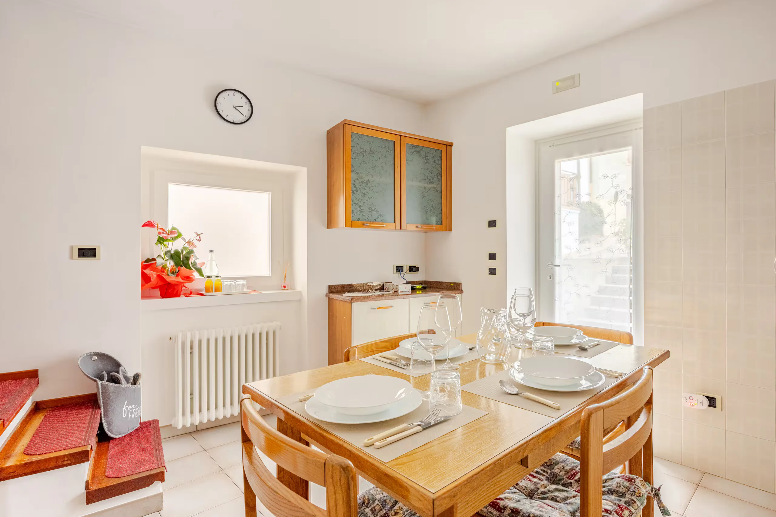 Tavolo in legno apparecchiato per quattro accanto alla porta finestra, con pensili e credenza